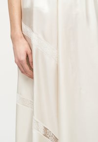 Αριστερό χέρι ακουμπισμένο δίπλα σε ροή από κρεμ σατινέ ύφασμα διακοσμημένο με διαγώνιες ημιδιαφανείς λωρίδες δαντέλας.