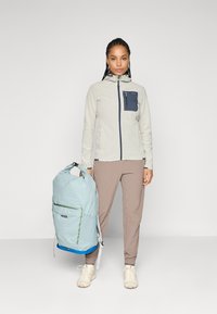 Frau trägt eine hellgemusterte Jacke mit Reißverschluss und beige Hosen, hält einen großen hellblauen Rucksack und steht vor einem schlichten Hintergrund.