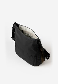 Schwarze Umhängetasche mit verstellbarem Riemen, offener Reißverschluss zeigt beiges Innenfutter mit Reißverschluss- und Elastikfächern, vor einem weißen Hintergrund.