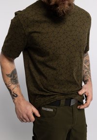 Olijfgroen T-shirt met een geometrisch zwart patroon, korte mouwen, gedragen met donkergroene broeken met een merkband en zakken.