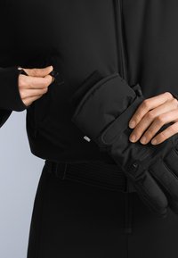 Ręce trzymające czarną zimową rękawiczkę i rozpinające kieszeń na czarnej kurtce na neutralnym tle.