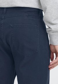 Temno mornarsko modre bombažne hlače z zadnjim žepom v subtilni, špičasti obliki. Tkanina ima teksturiran videz in klasičen kroj.