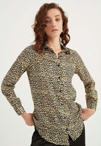 Leopardprint knap skjorte i beige med sorte mønstre, lange ærmer og rundet kant. Glat stof med klassisk krave.