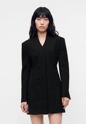 Un mannequin aux longs cheveux foncés se tient face à l’objectif, vêtu d’une robe blazer noire à manches longues, sur un fond blanc uni.