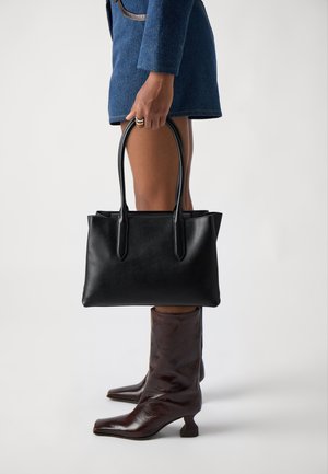 Borsa a tracolla nera in pelle con due manici, forma rettangolare, texture liscia. Modello che indossa stivali in pelle marrone con punta quadrata.