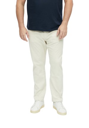 Mann trägt cremefarbene Hosen, weiße Sneaker und ein dunkel marineblaues Hemd und steht vor einem weißen Hintergrund.