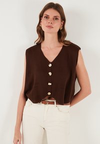 Brązowy dziergany top bez rękawów z dekoltem w kształcie litery V, pięcioma dużymi guzikami i subtelnymi bocznymi kieszeniami, zestawiony z białymi spodniami i brązowym paskiem.