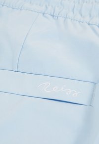 Nahaufnahme eines hellblauen Stoffes mit gesammeltem elastischem Bund und weiß gesticktem „Reiss“-Logo auf angenähter Borte.