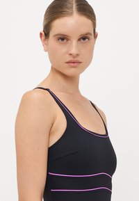 Woman with slicked-back hair wearing a black tank top with thin purple piping, facing forward with a neutral expression, white background.