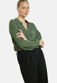Moteris su šviesia oda ir šviesiais plaukais, susirišusiais į žemą kuodą, dėvi žalią bluzą su nėriniuota detale ir juodas aukštai padidintas kelnes, rankas sukryžiavusi.
