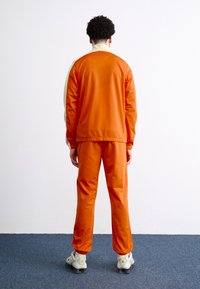 Πορτοκαλί φόρμα με αντίθετο κρεμ γιακά, ελαστικούς καρπούς και πέφτει χαλαρά πάνω στο σώμα. Συνδυάζεται με γκρί και λευκά αθλητικά παπούτσια.