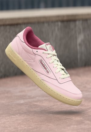 Baskets Reebok roses avec une semelle beige, des lacets blancs et un logo bordeaux, flottant au-dessus d'une surface en béton rose avec un fond gris.