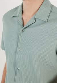 Jasnozielona koszula z krótkim rękawem o prążkowanej fakturze, z kołnierzykiem i dwoma guzikami z przodu.