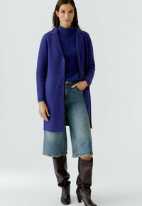 Lange paarse jas van textuurstof, met een dubbel borst ontwerp, gecombineerd met een blauwe coltrui en wijde denim shorts. Bruine kniehoog laarzen.