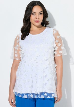 Kobieta o ciemnych włosach ubrana w biały top z przezroczystymi rękawami zdobionymi kwiatowymi aplikacjami oraz niebieskie spodnie, stojąca na białym tle.