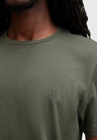 T-shirt in cotone verde oliva con colletto rotondo e maniche corte. Presenta un piccolo design di teschio ricamato sul lato sinistro del petto.