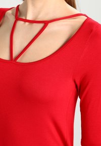 Top rojo ajustado con escote redondo y detalles de tiras entrecruzadas. La tela parece suave y elástica, proporcionando una silueta ceñida.