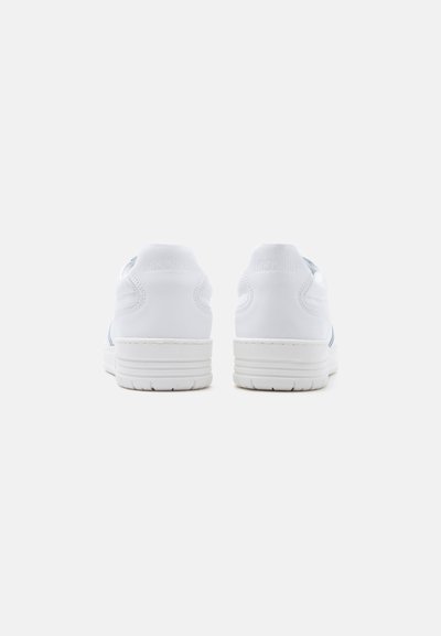 Een paar witte sneakers gezien van de achterkant met dikke zolen en minimale stikseldetails op een effen lichte achtergrond.