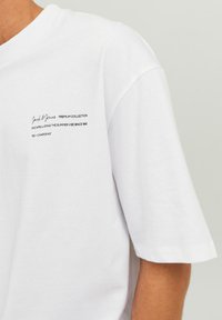 Hvid bomulds t-shirt med rund hals og korte ærmer. Har sort tekst på venstre side, der fremhæver brand- og kollektionsdetaljer.