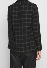 Svart blazer med vit rutmönster, lättviktsmaterial, slagkrage och skräddarsydd passform. Har en slits i ryggen och rena sömmar.