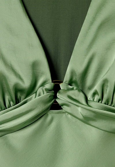 Robe en satin vert avec un décolleté plongeant, un tissu froncé et un accent circulaire en or à la taille. Texture lisse et design élégant.