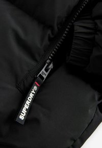 Juodas pūkinis švarkas, medžiagos detalus vaizdas, kuriame matyti užtrauktukas ir etiketė su užrašu "SUPERDRY". Medžiaga atrodo lygi su siūlėmis.