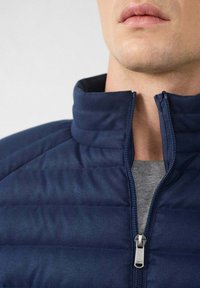 Hombre con una chaqueta acolchada azul marino con un cuello alto parcialmente abrochado y una camisa gris debajo.