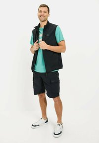Sort vest over en teal t-shirt, parret med sorte shorts. Hvide sneakers med marinefarvede detaljer. Glat stof, afslappet udendørs tøj.