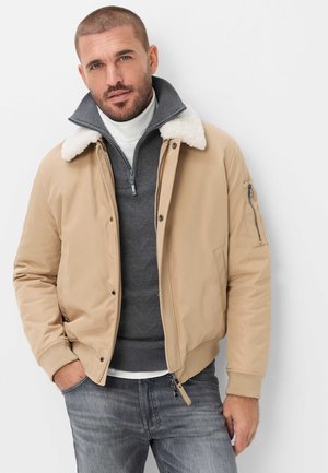 Mand iført beige jakke med hvidt lammeskindskrave, grå lynlås sweater, hvid højhalset trøje og grå jeans står mod hvid baggrund.