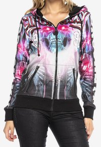 Kvinde iført en lynlås hoodie med et farverigt skelet, lyn og mekanisk design, parret med sorte teksturerede bukser.