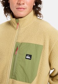 Fleece jacket in beige with a green chest pocket. Features a zip closure with red accents and a logo patch. Textured fabric adds warmth.