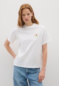 Joven con cabello rojo que lleva una camiseta blanca con un pequeño oso de peluche y bordado de laurel, combinada con jeans de un azul claro, frente a un fondo liso.