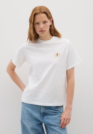 Jeune femme aux cheveux roux portant un t-shirt blanc avec une petite broderie d'ours en peluche et de laurier, associé à un jean bleu clair, sur fond uni.