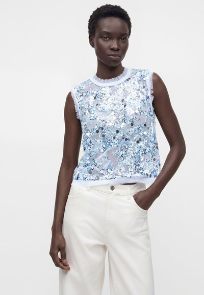 Γυναίκα που φοράει ένα αμάνικο ανοιχτό μπλε τοπ με παγιέτες και λευκό παντελόνι, με το ένα χέρι στην τσέπη, στέκεται μπροστά σε απλό φόντο.
