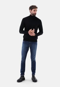 Schwarzer Rollkragenpullover in einem taillierten Design, kombiniert mit Jeans in mittlerer Waschung. Das Model trägt schwarze Knöchelstiefel und steht vor einem schlichten Hintergrund.