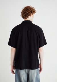 Camisa de lino negra de manga corta con cuello, corte relajado y dobladillo recto, llevada sobre unos jeans de mezclilla azul claro. Diseño sencillo, sin patrones.