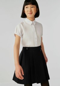 Camicia bianca a maniche corte con bottoni e cuciture decorative, abbinata a una gonna nera plissettata con zip laterale e dettaglio con bottone.