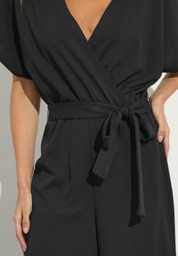 Abito tuta nero con maniche corte e cintura a fiocco in vita. Il tessuto appare liscio con un lieve drappeggio e dettagli minimi.