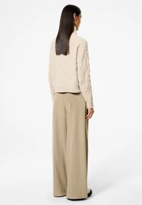 Maglione beige lavorato a maglia con texture a diamante e colletto alto. Abbinato a pantaloni larghi di un beige chiaro. Scarpe nere slip-on in vista.