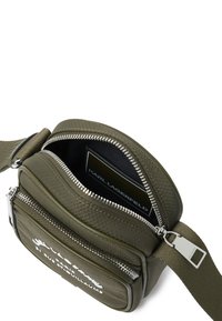 Borsa a tracolla verde oliva in materiale testurizzato, con zip argentata, dettaglio del logo e tracolla regolabile, parzialmente aperta per rivelare l'interno.