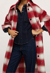 Červenohnědý a bílý kostkovaný oversized kabát s knoflíky, nošený přes tmavou džínovou košili a kalhoty. Tkanina vypadá měkce a teple.