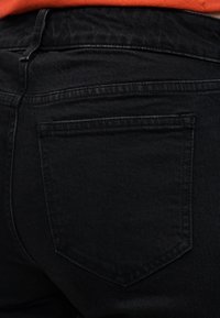 Κοντινό πλάνο ενός ατόμου που φοράει μαύρο τζιν τζιν με την πίσω τσέπη και μέρος ενός πορτοκαλί πουκάμισου στη μέση.