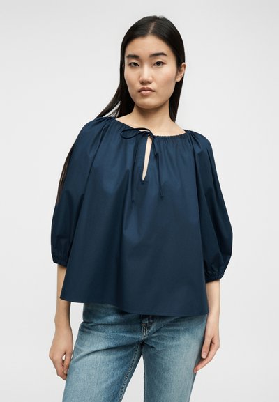 Mladá žena s dlhými tmavými vlasmi, ktorá má na sebe voľnú námornícku blúzku s nafúknutými rukávmi a výstrihom na zapínanie, v kombinácii so svetlomodrými džínsami.