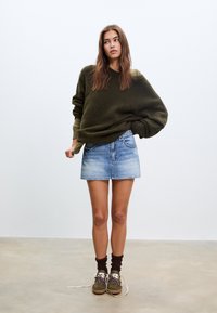 Pull oversize vert olive, mini-jupe en denim bleu clair, chaussettes hautes marron jusqu'aux genoux, et chaussures vertes avec lacets blancs et accents à motifs.