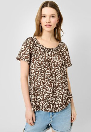 Jeune femme aux longs cheveux portant un chemisier marron à manches courtes à motif floral et un short en jean bleu clair sur un fond uni.