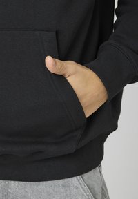 Hoodie en coton noir avec une poche kangourou, des poignets et un ourlet côtelés. Le tissu semble doux avec une texture lisse. Pantalons de survêtement gris visibles.