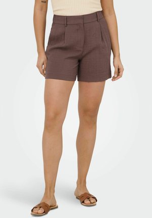 Kobieta w brązowych, dopasowanych szortach z wysokim stanem, zestawionych z jasnobrązowymi sandałami na płaskim obcasie i beżowym topem bez rękawów.