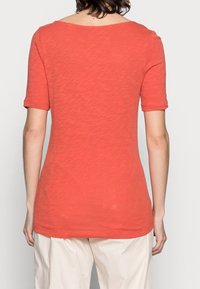 T-shirt a maniche corte, con collo rotondo, di colore corallo, realizzato in tessuto leggero con una finitura leggermente strutturata, indossato con pantaloni di colore crema.