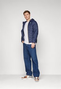 Doudoune marine avec capuche, design matelassé, fermeture éclair dorée, t-shirt blanc en dessous, jean bleu ample, et baskets avec accents bruns.
