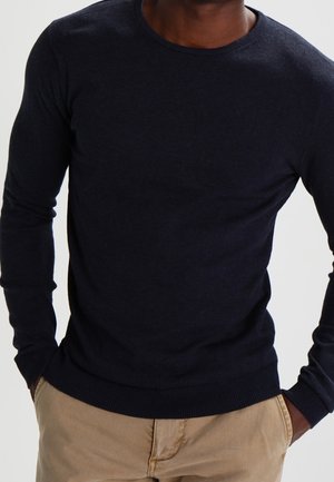 Maglione a girocollo di colore blu navy realizzato in tessuto a maglia, con maniche lunghe e polsini a costine, abbinato a pantaloni chiari.